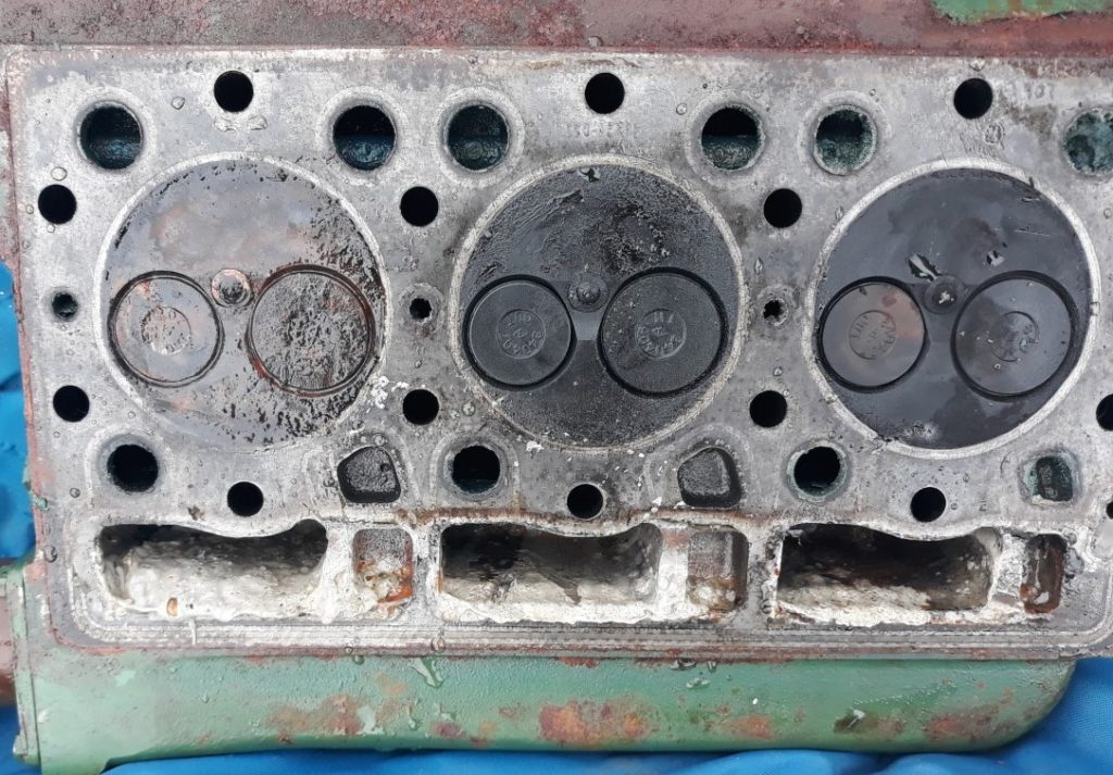 image of The underneath of the engine head showing the top of the three cylinders. Each has two valves-one opens to let air in and the other lets exhaust gases out. Seawater has got into the left hand one. Underneath there is a thick off white cream coloured mixture of oil and seawater.